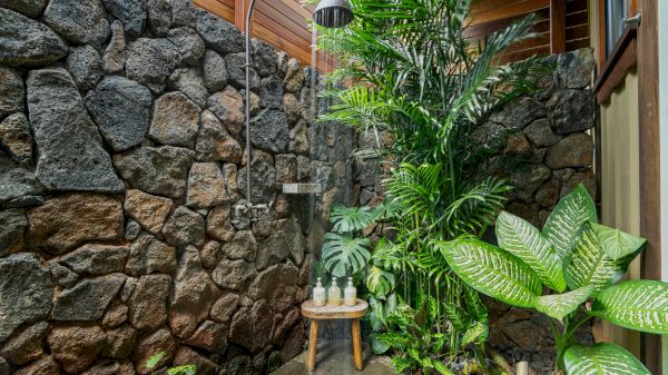 An outdoor shower with stone walls, lush green plants, and a wooden stool holding toiletries under a rainfall showerhead.
