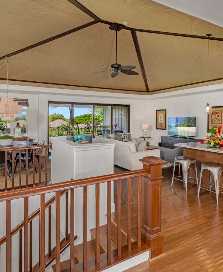 A bright, open-concept living area with a kitchen bar, island seating, a dining table, white chairs, and a cozy lounge, overlooking a sunny backyard.