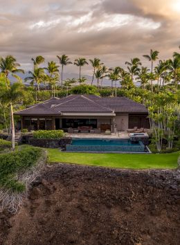 This image shows a luxurious house with a pool surrounded by palm trees, set on a cliffside with a scenic backdrop and cloudy sky.
