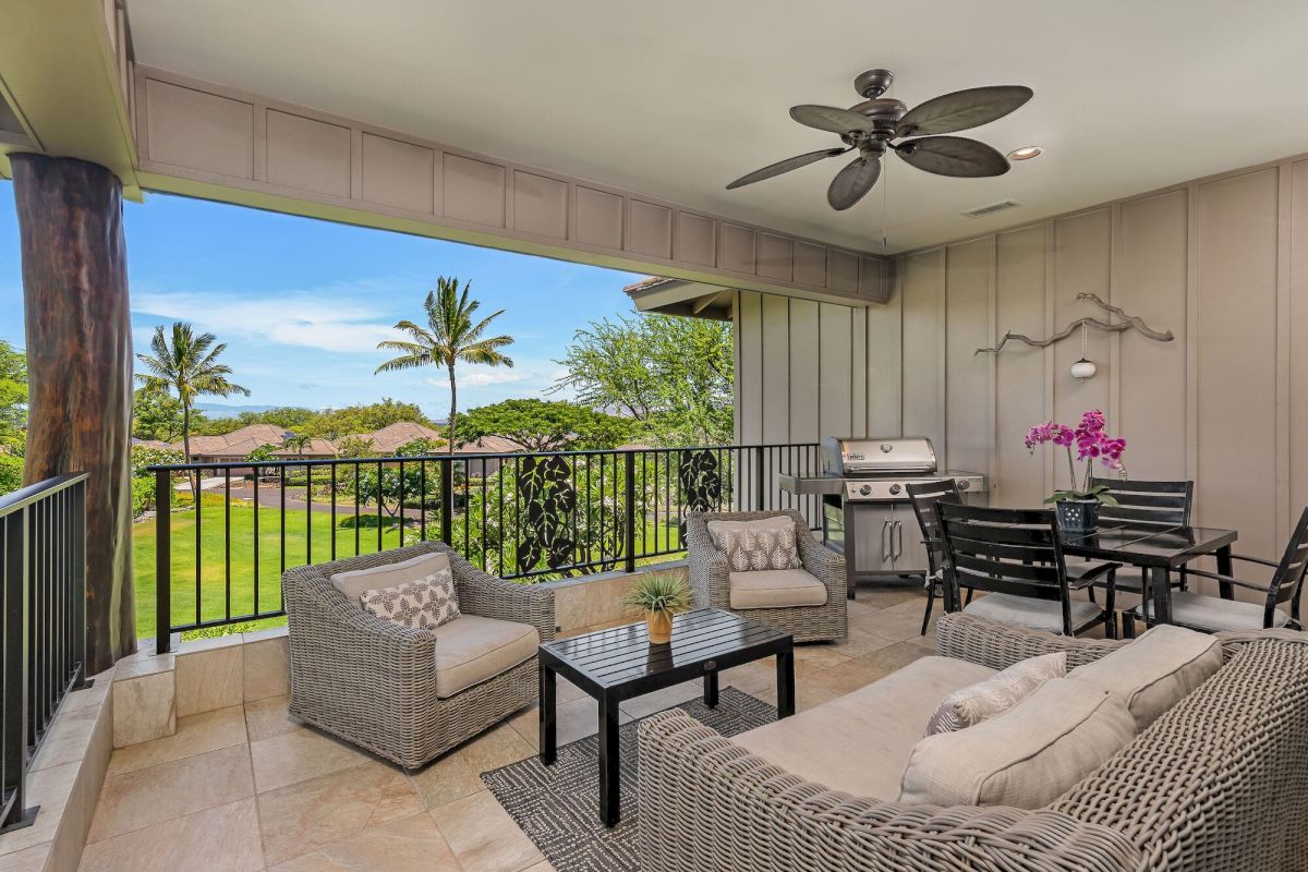 A covered outdoor patio features wicker furniture, a dining table, grill, and ceiling fan, overlooking a lush landscape with palm trees.