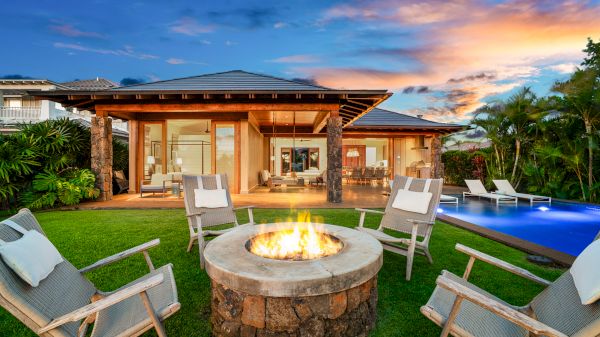 A modern house with a fire pit, surrounded by chairs, and a pool in the backyard under a colorful sunset sky.