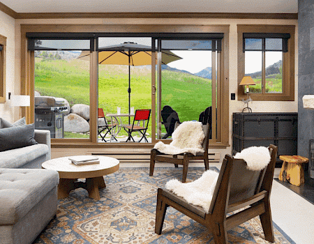 A cozy living room with a gray sofa, patterned rug, fireplace, and large windows opening to a patio with green hills outside.