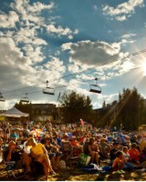 A large crowd sits outdoors under a partly cloudy sky, with ski lifts visible overhead and the sun shining brightly in the afternoon.