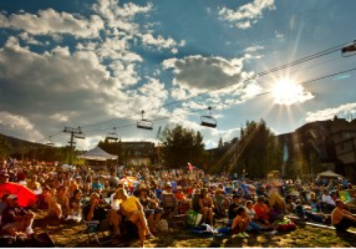 A large crowd sits outdoors under a partly cloudy sky, with ski lifts visible overhead and the sun shining brightly in the afternoon.