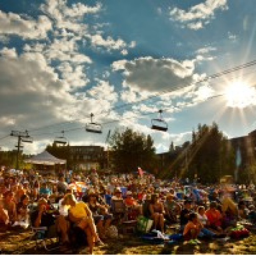 A large crowd sits outdoors under a partly cloudy sky, with ski lifts visible overhead and the sun shining brightly in the afternoon.