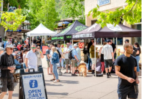 A bustling outdoor scene with people visiting market stalls under tents. A sign indicates "OPEN DAILY" for an information center or shop.