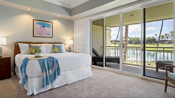 A bright bedroom with a large bed, blue throw, window view of palm trees and water, and a hat on the bed, creating a serene atmosphere.