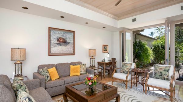 A bright, spacious living room with a gray sofa, floral chairs, yellow pillows, a wooden coffee table, and large sliding doors opening to a green outdoor area.