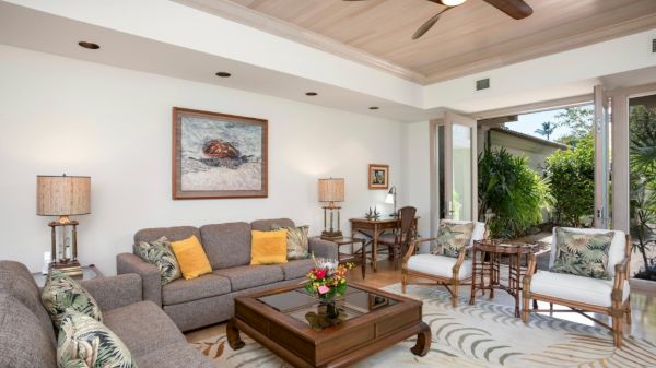 A cozy living room with gray sofas, a wooden coffee table, tropical cushions, lamps, artwork, and large windows opening to greenery.