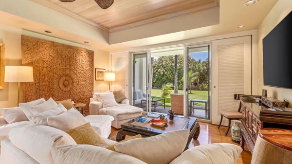 A bright, airy living room with white sofas, a wooden coffee table, warm lamps, a large screen, and sliding glass doors opening to a green garden.