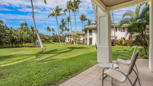 A sunny tropical resort lawn with palm trees, a covered patio, and patio chairs overlooking a courtyard between light-colored buildings.