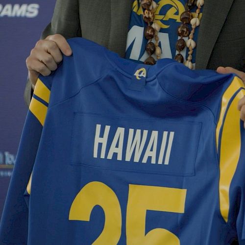 A person holds a blue sports jersey with "HAWAII" and the number 25 on it, likely from a team affiliated with Los Angeles.