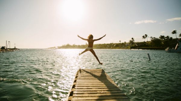 A person jumps on a dock over water with the sun setting in the background, creating a silhouette effect.