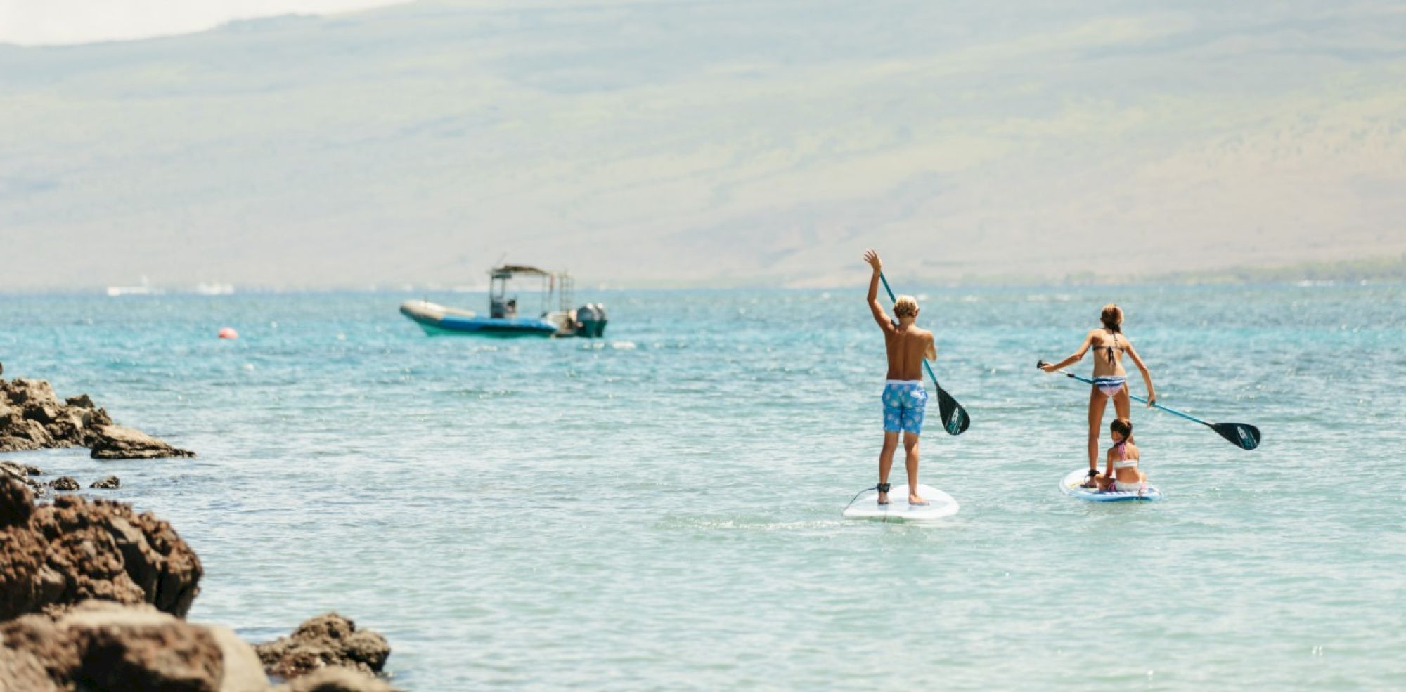 Two people paddleboarding on a calm ocean with a distant boat and rocky shoreline, enjoying a sunny day.