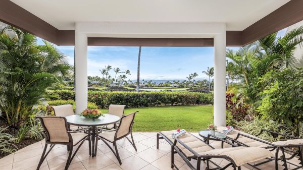 A covered patio with a round table and four chairs, facing a manicured lawn and distant palm trees in a tropical backyard.