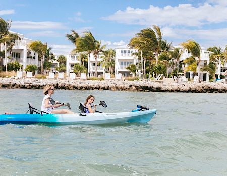 Two people paddle a small blue kayak near a sunny shoreline with palm trees and white buildings in the background.