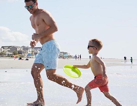 A man and child in swimsuits run along a beach, with the child holding a green frisbee. The sky is clear and the ocean is visible.