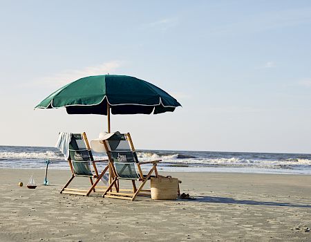 Two beach chairs under a green umbrella on a sandy shore, facing the sea with waves, and a basket nearby.