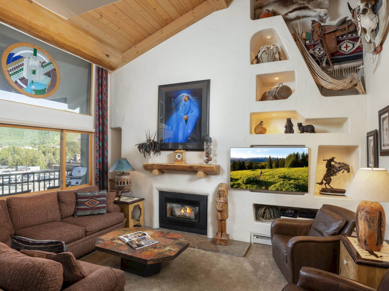 A cozy living room with a high ceiling, featuring a fireplace, eclectic art, and a large window offering scenic views of greenery outside.