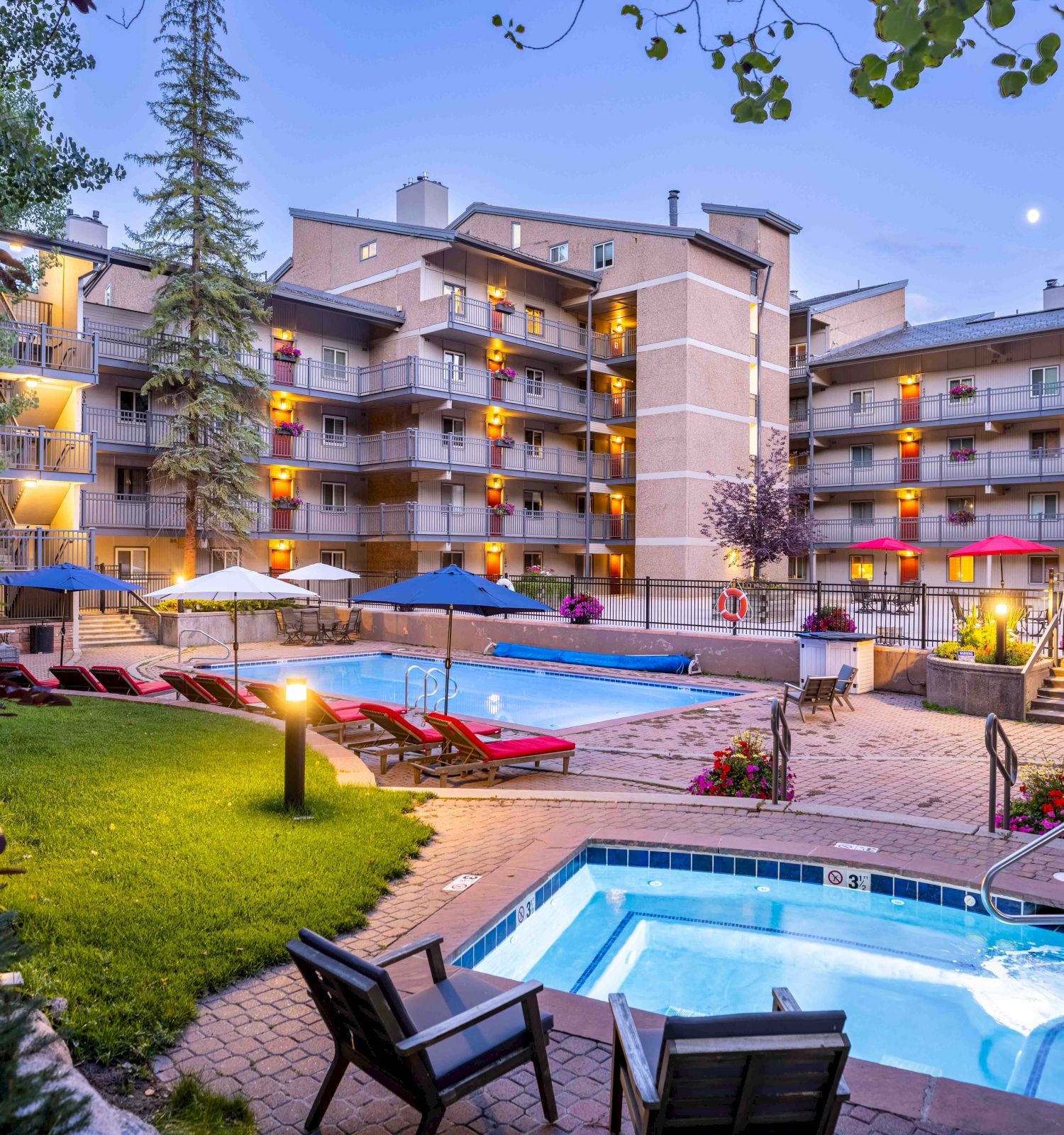 The image shows a hotel courtyard with a pool, hot tub, red lounge chairs, and a building with balconies surrounded by trees at twilight.