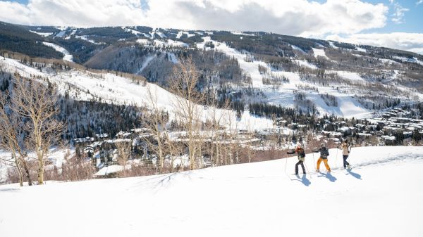 A snow-covered mountain landscape with three people hiking, clear skies, and distant views of a town nestled in the valley below.