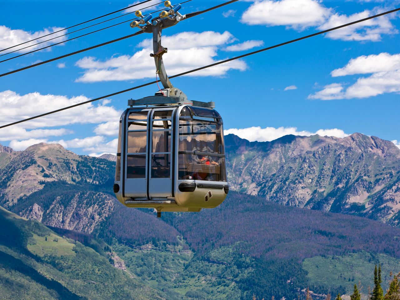 A cable car is suspended in the air against a backdrop of scenic mountains and a clear blue sky with scattered clouds.