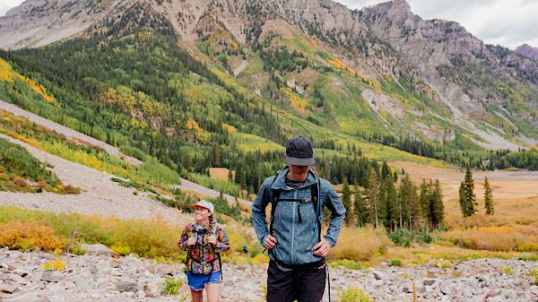 Two people hiking in a scenic mountainous area with lush greenery and colorful autumn foliage.