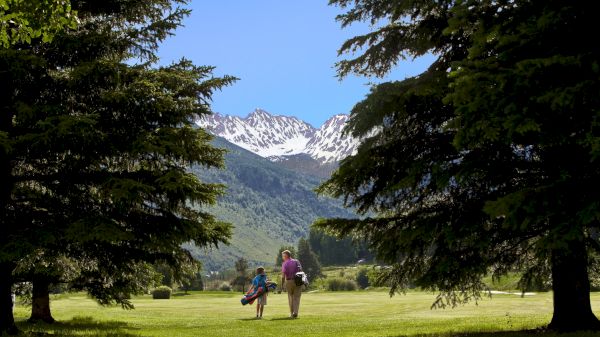 Two people walking on a lush golf course, surrounded by trees, with a scenic view of snow-capped mountains in the background.