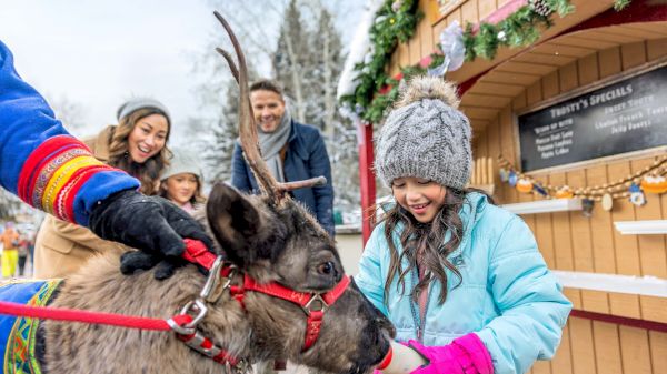 A young girl in winter clothing feeds a reindeer while a family looks on near a festive booth adorned with holiday decorations.