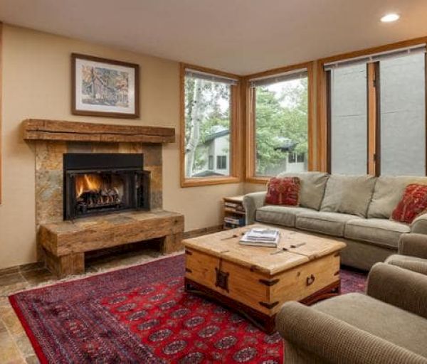 A cozy living room features a fireplace, two sofas, a TV, and a wooden coffee table on a red rug, with windows bringing in natural light.