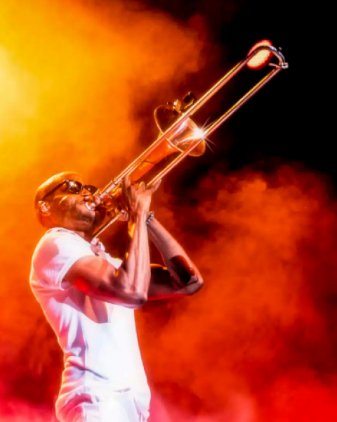 A musician passionately plays a brass trombone on a smoky, fiery stage, shouting or singing into the instrument, bathed in orange light.