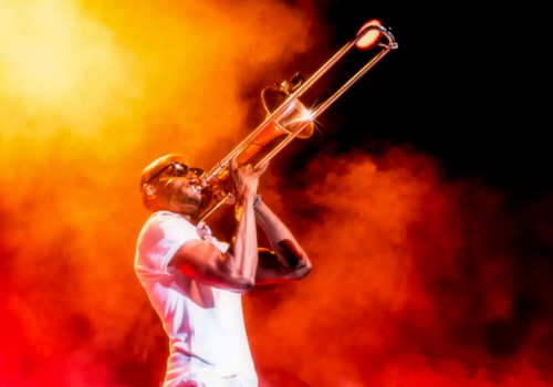 A musician passionately plays a brass trombone on a smoky, fiery stage, shouting or singing into the instrument, bathed in orange light.