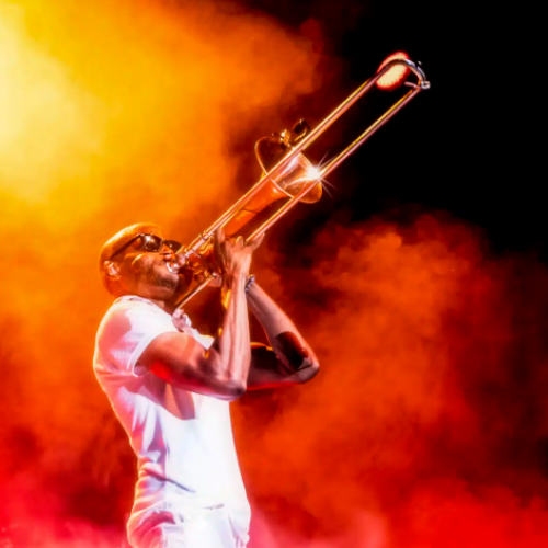 A musician passionately plays a brass trombone on a smoky, fiery stage, shouting or singing into the instrument, bathed in orange light.