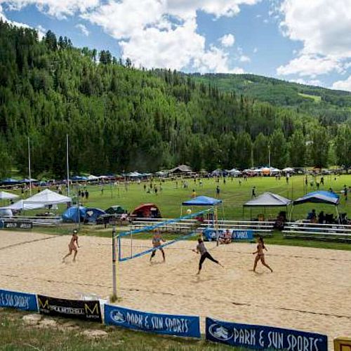 The image shows a beach volleyball game happening outdoors, surrounded by a lush green landscape and tents.