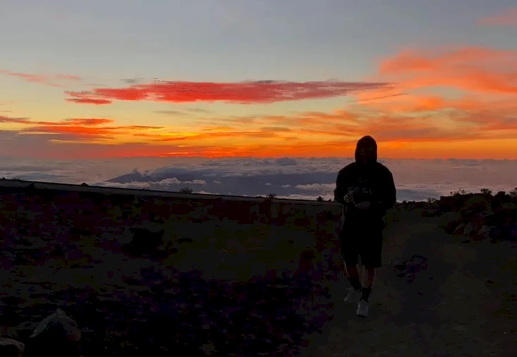 A person stands silhouetted against a vibrant sunset sky with clouds and mountains in the background.
