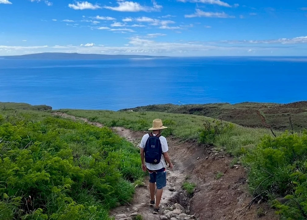 A person with a hat and backpack walks on a trail through greenery, overlooking a vast ocean under a blue sky with clouds.