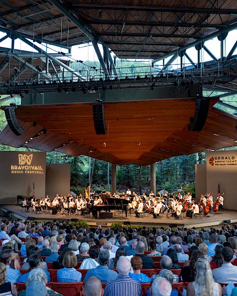 An outdoor concert with an orchestra performing under a large canopy, watched by a seated audience in a scenic, wooded setting.