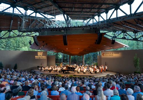 An outdoor concert with an orchestra performing under a large canopy, watched by a seated audience in a scenic, wooded setting.