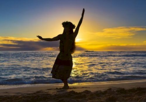 A person performs a hula dance on the beach at sunset, silhouetted against the sea and colorful sky.