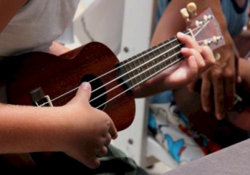 Two people are holding a ukulele, one playing it while the other assists, in a casual indoor setting.