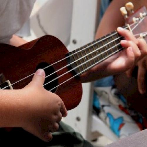Two people are holding a ukulele, one playing it while the other assists, in a casual indoor setting.