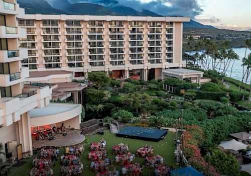 A hotel by the ocean with a lush garden, outdoor seating, and a gathering area under a clear sky with mountains in the background.