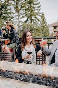 People are enjoying drinks around a fire pit with trees in the background, creating a cozy outdoor setting.