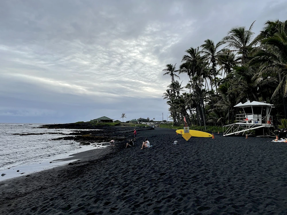 A black-sand beach with palm trees, a lifeguard tower, and a few people near the shore at sunset, on a calm coastal scene.
