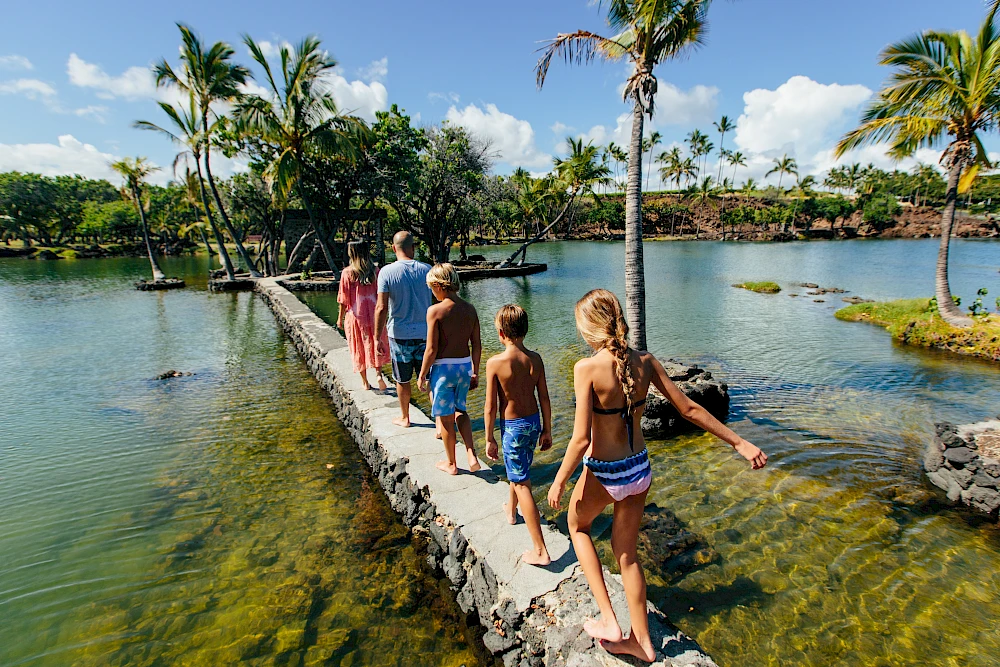 A group of people walks along a narrow stone bridge over clear tropical water, palm trees sway, and a sunny blue sky frames a tropical scene.