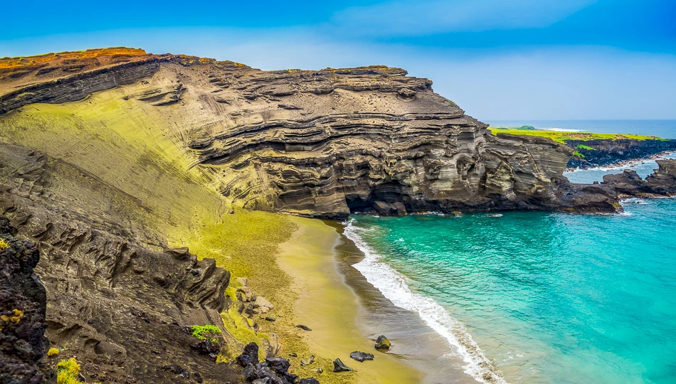 A rugged coastal landscape with layered cliffs, a yellowish-sand beach, and turquoise waves meeting the rocky shore at bright blue sky.