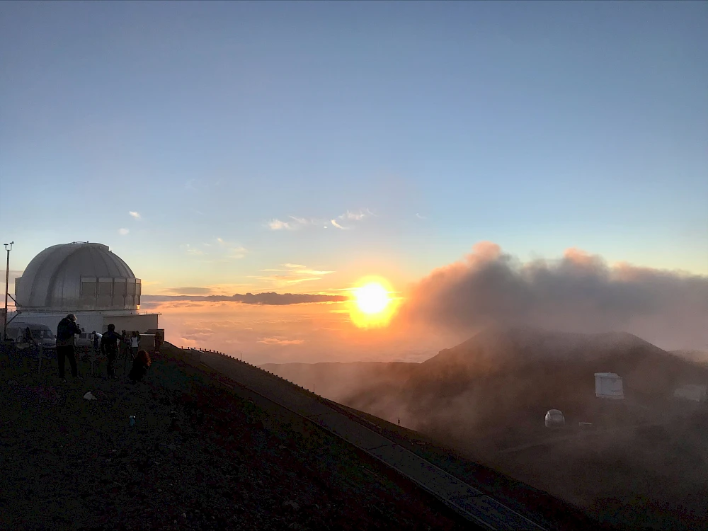 A sunset over a hilltop observatory with a dome, people near the rim, and wispy clouds illuminated by the warm glow of the sun.
