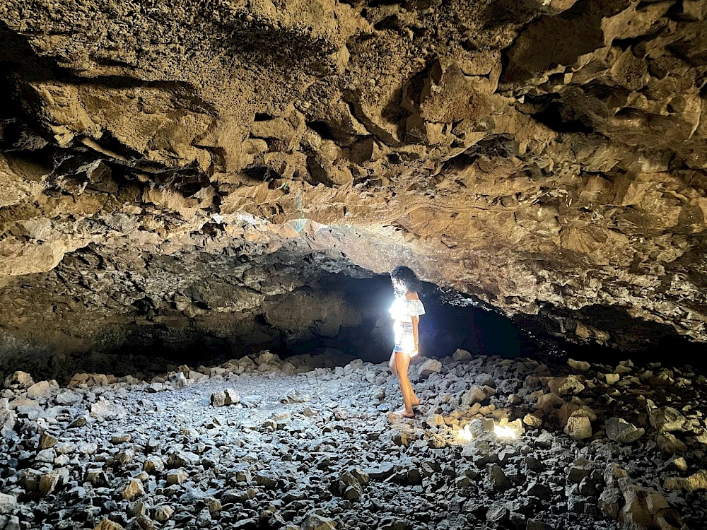 A person stands inside a rocky cave with rough walls and a bright light shining, dust and debris on the ground, natural cave interior.