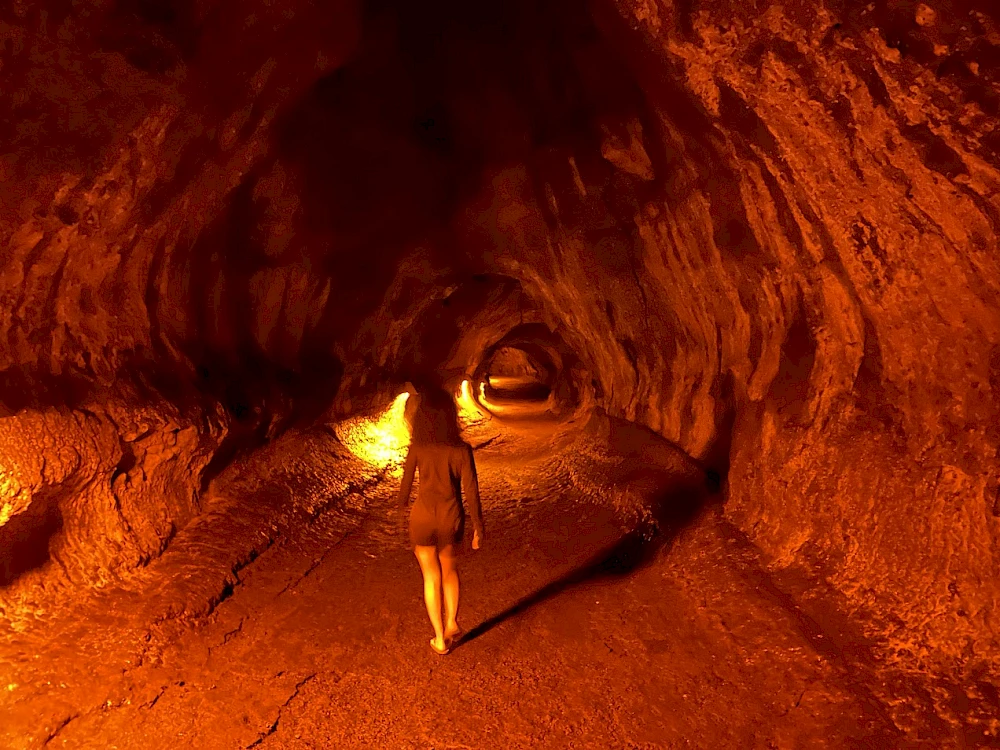 A person walks through a rocky tunnel or cave, glowing orange-lit walls and a rough, curved path ahead, ending at a dim exit.