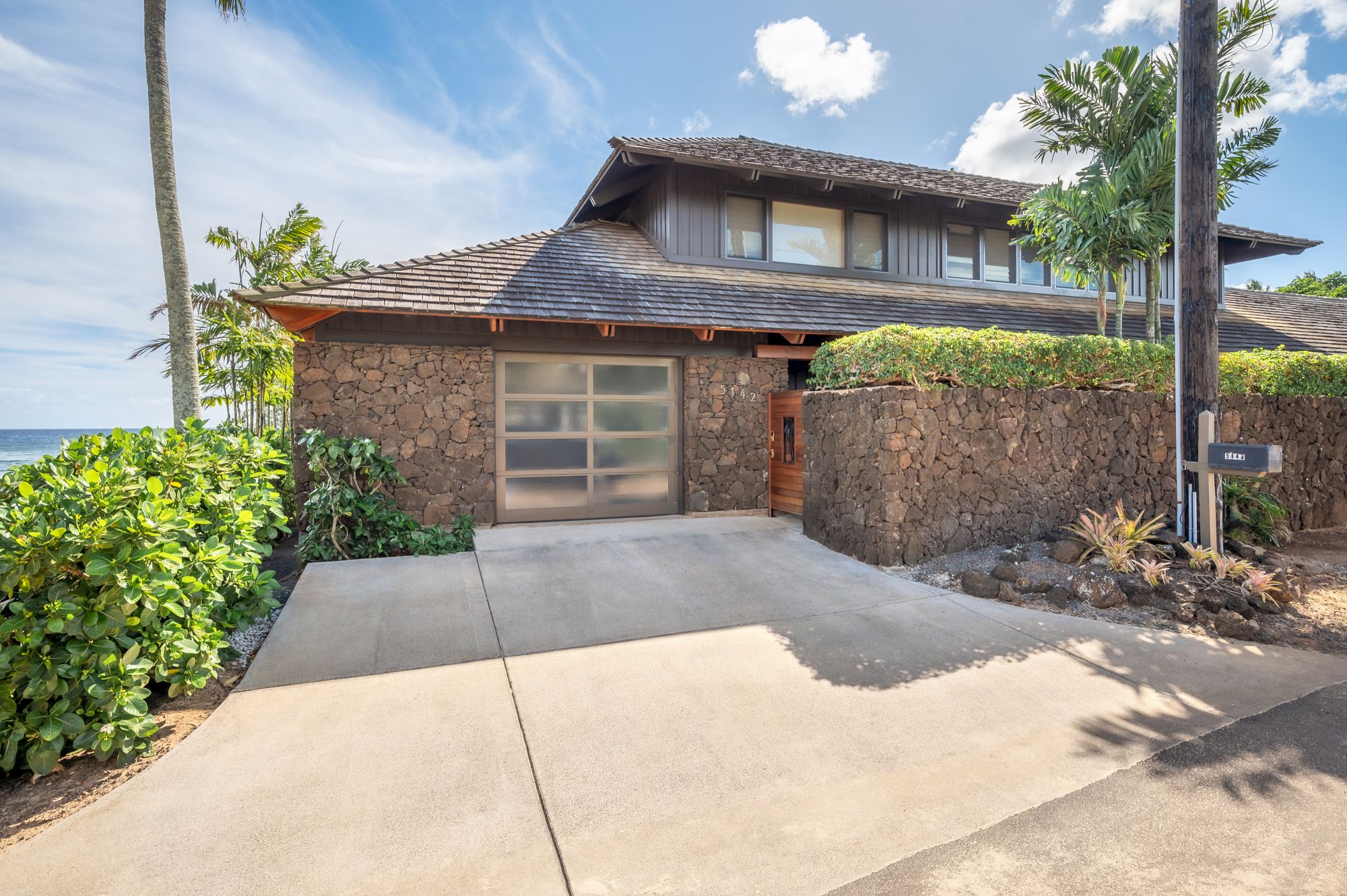 A modern house with a stone facade, large garage door, and surrounded by greenery, set near the ocean under a clear blue sky.